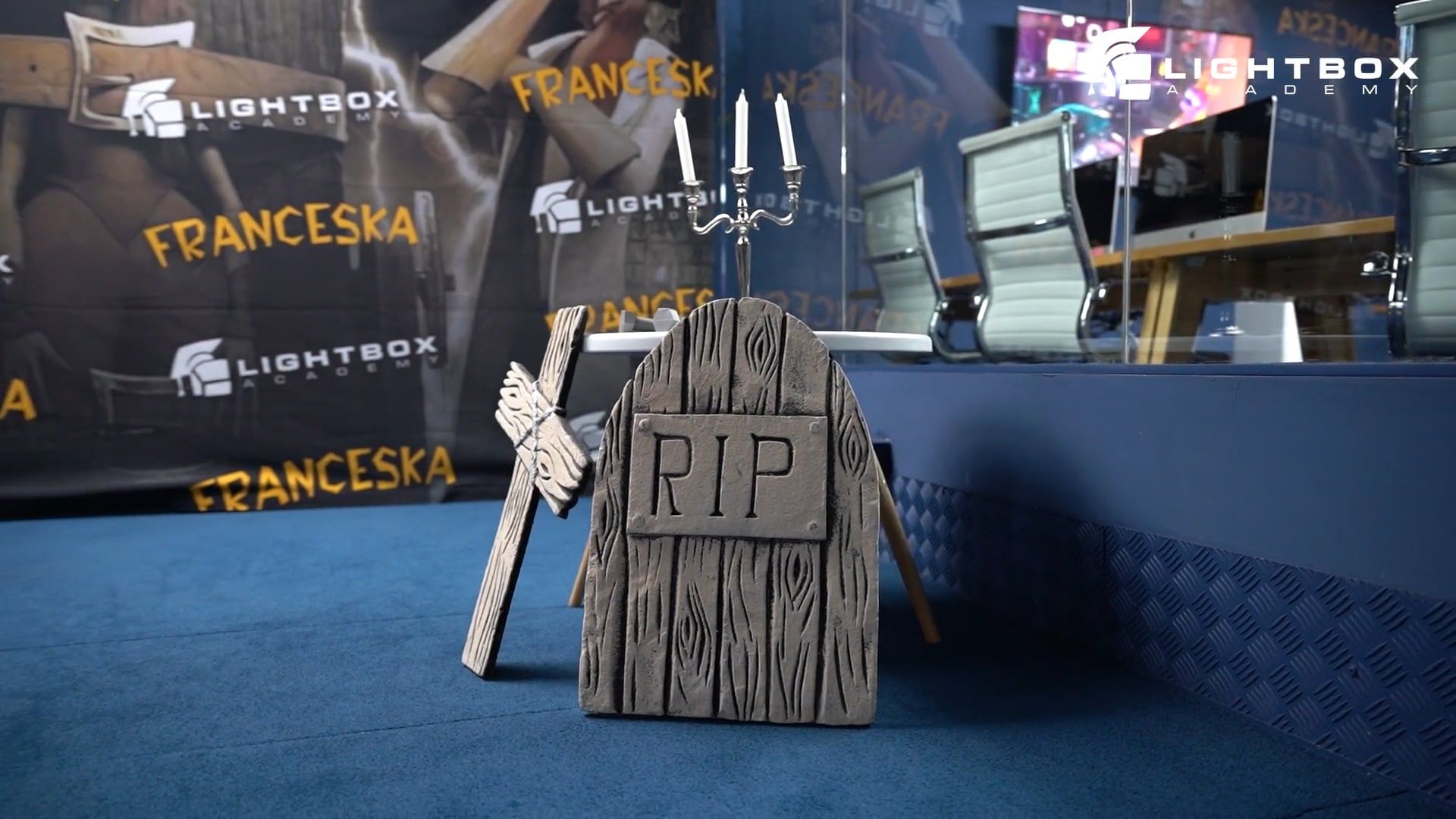 Dispositivo decorativo en forma de lápida con inscripción “RIP” sobre suelo azul, con mobiliario y pantallas de Lightbox Academy al fondo.