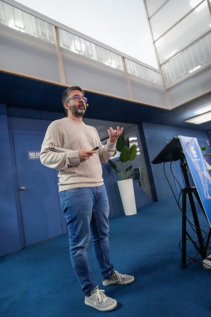 Josep Gatell con jersey claro y vaqueros, sosteniendo utensilios, en actitud explicativa junto a atril en sala azul.
