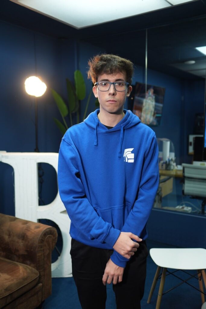 Pastor-4.jpg Fotografía vertical de cuerpo casi entero de un joven con sudadera azul y gafas, de pie junto a sofá y mesas en Lightbox Academy.