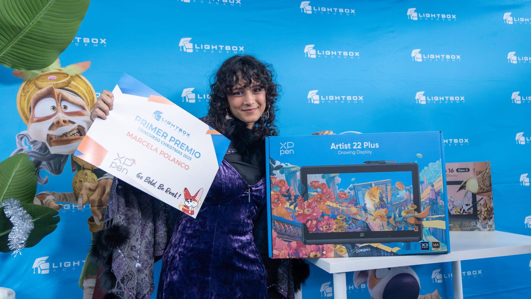Marcela Polanco con vestido morado y cabello rizado sostiene con orgullo un diploma de "Primer Premio" y una caja grande de XP-pen Artist 22 Plus.