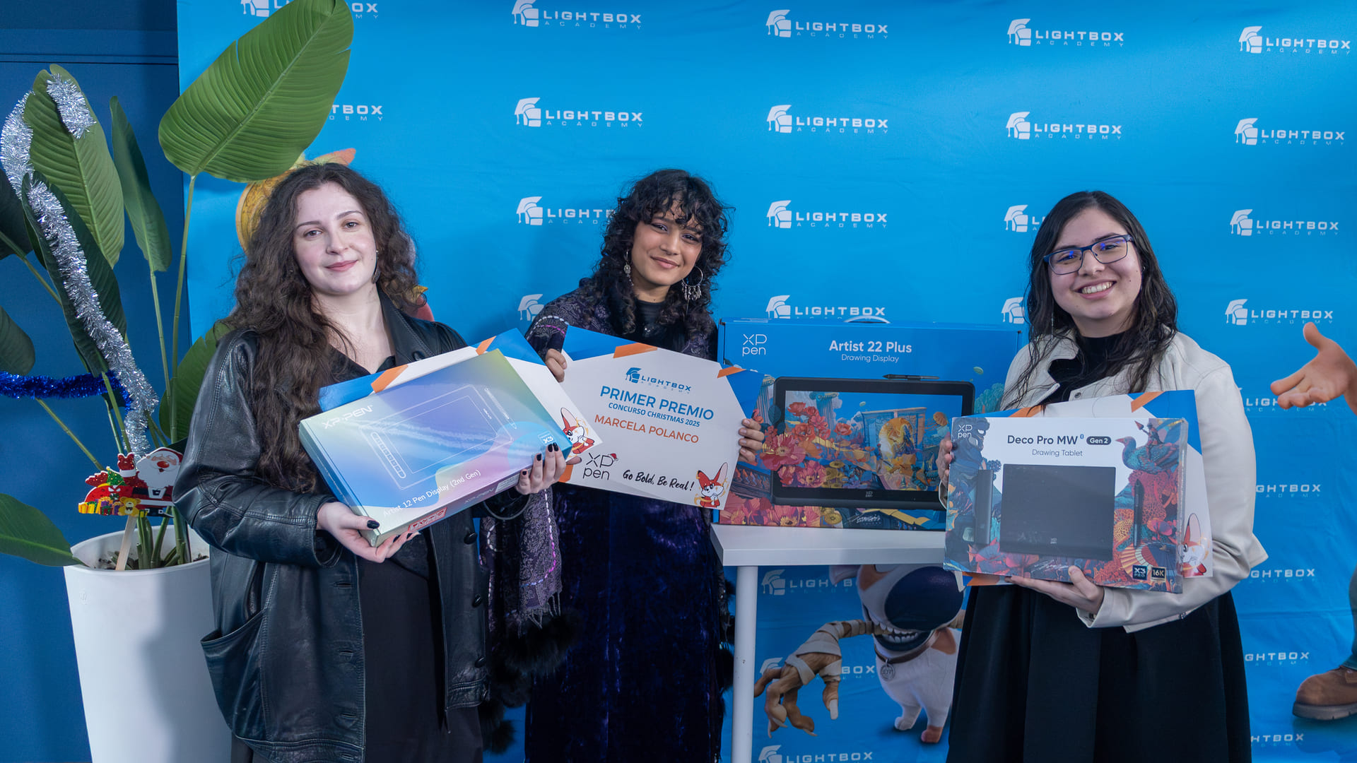 Las tres ganadoras alumnas posan juntas sosteniendo sus respectivos premios y diplomas frente a un mural azul con logotipos de Lightbox Academy.
