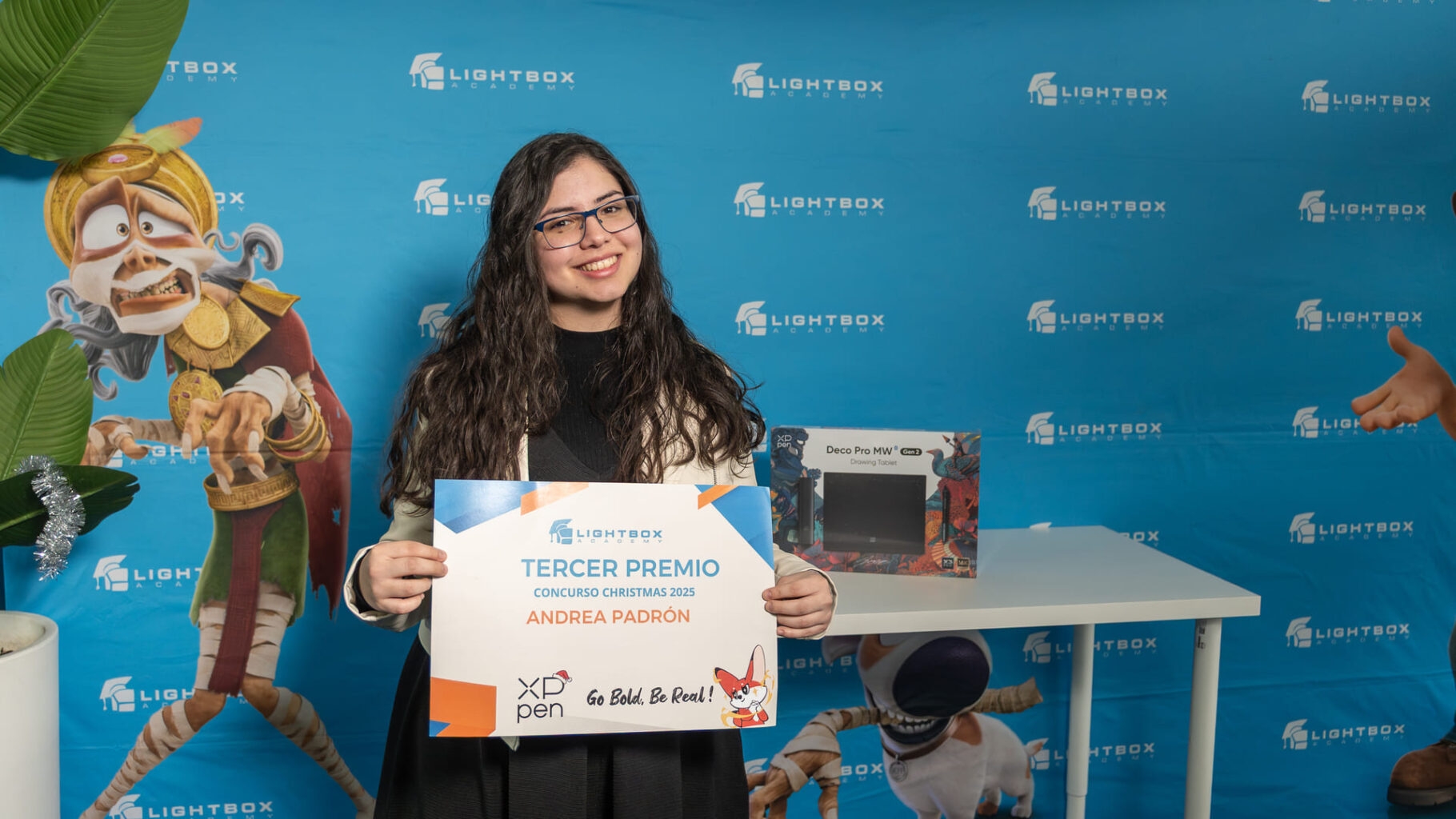 La alumna Andrea Padrón, con gafas y chaqueta blanca sonríe mientras sostiene un diploma de "Tercer Premio" del Concurso Christmas 2025 de Lightbox Academy.