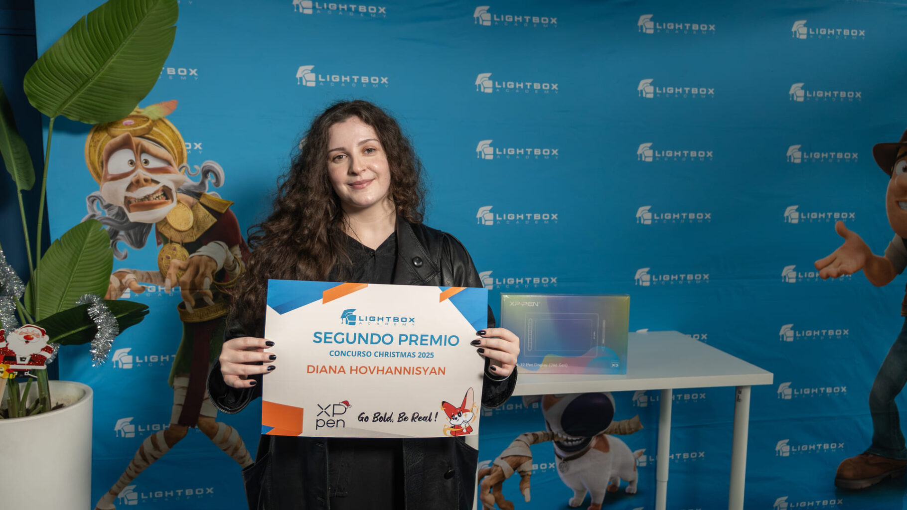 Diana Hovhannisyan con cabello largo y oscuro sostiene un diploma que indica "Segundo Premio" del Concurso Christmas 2025 frente a una mesa con una tableta gráfica.
