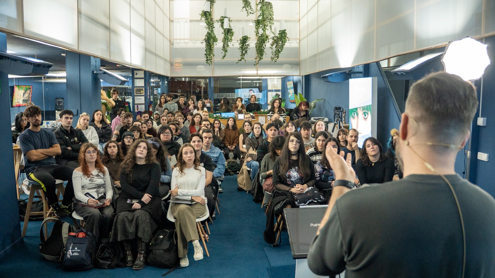 Una toma desde atrás del presentador gesticulando hacia una audiencia atenta sentada en un auditorio moderno con paredes azules y plantas colgantes.