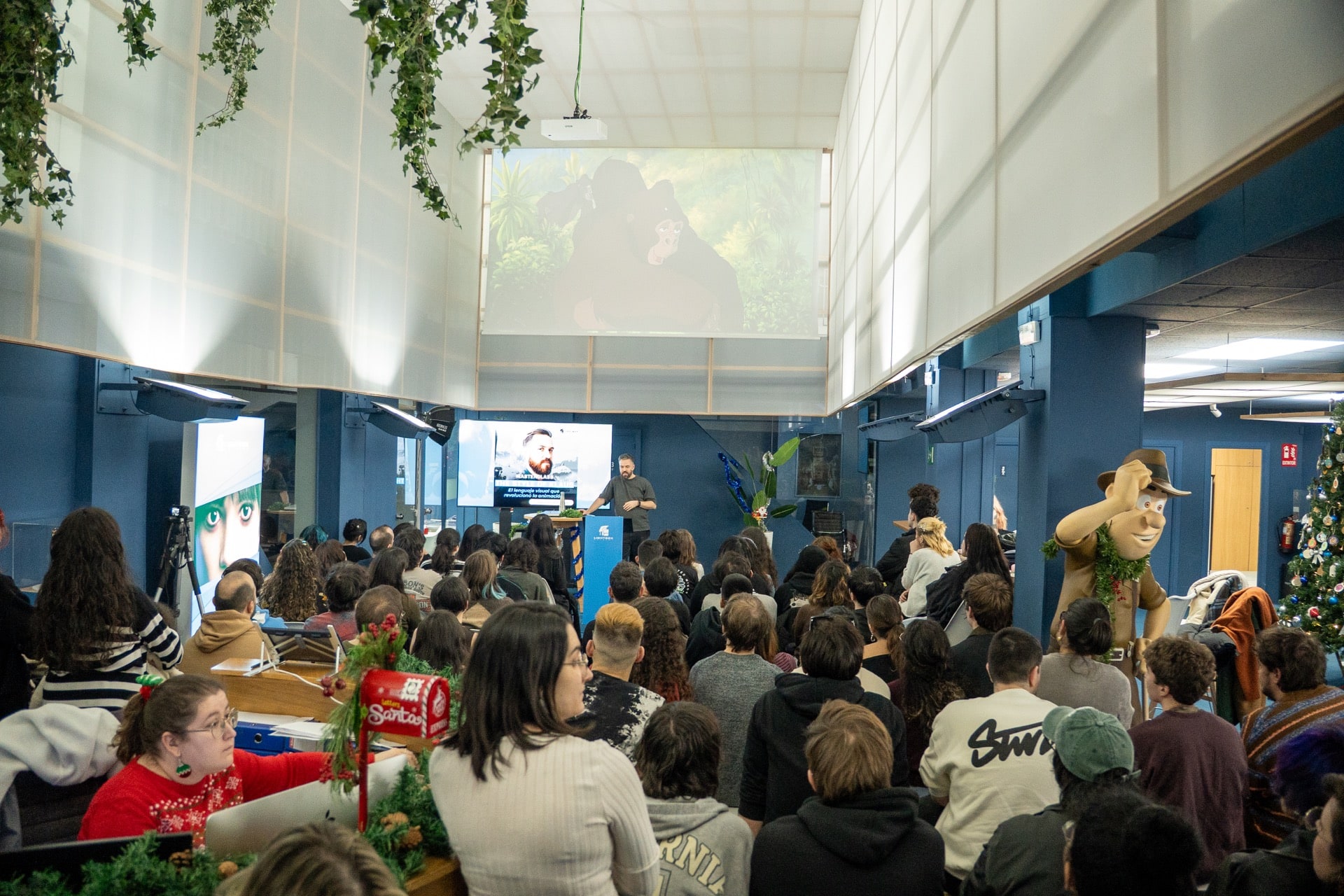 Vista amplia de una sala llena de estudiantes mirando hacia una pantalla gigante que proyecta una escena de animación con un gorila, mientras Szymon Biernacki habla desde un podio.