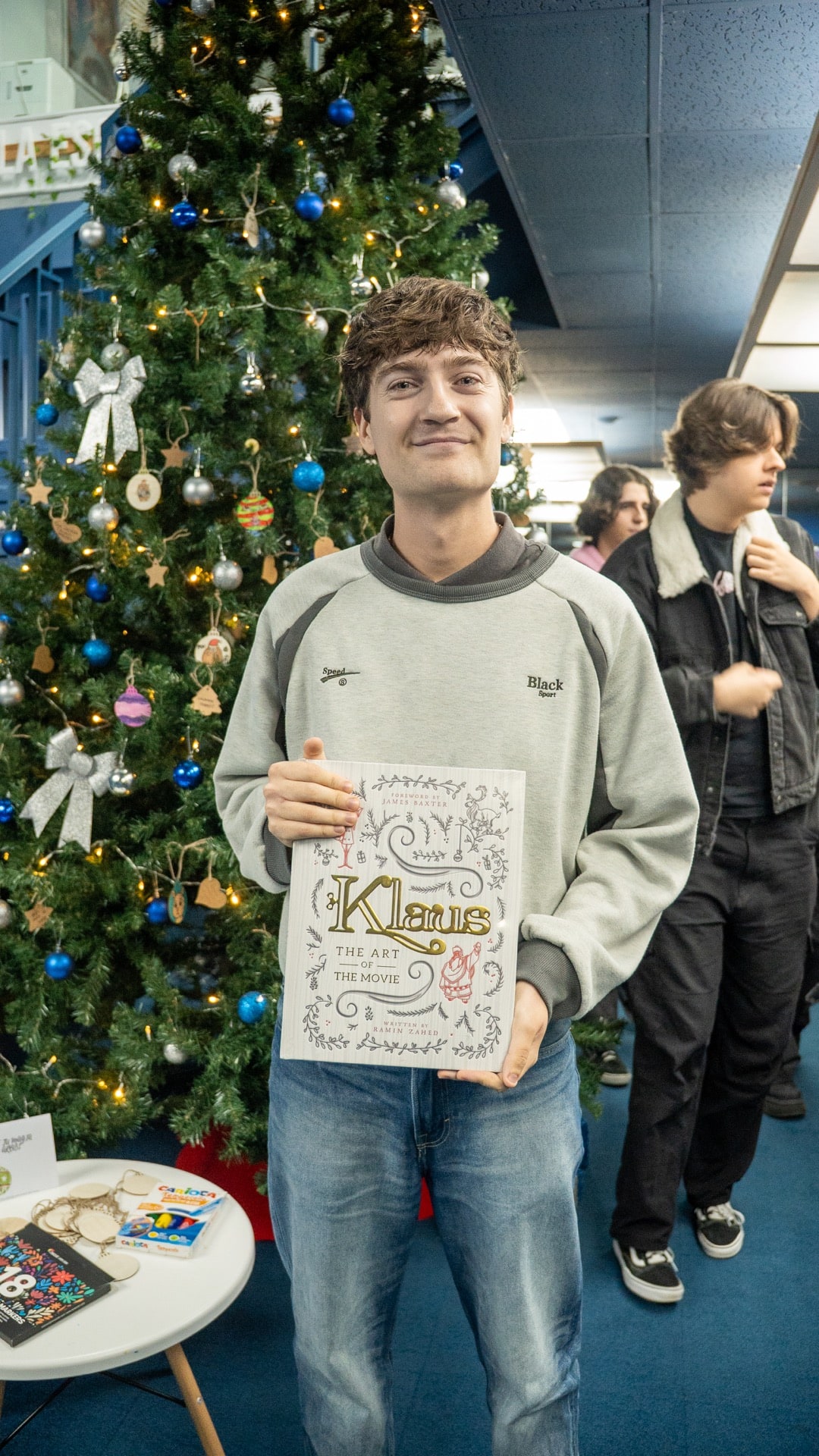 Estudiante posando frente a un árbol de Navidad sujetando el libro ilustrado “Klaus: The Art of the Movie” en Lightbox Academy.