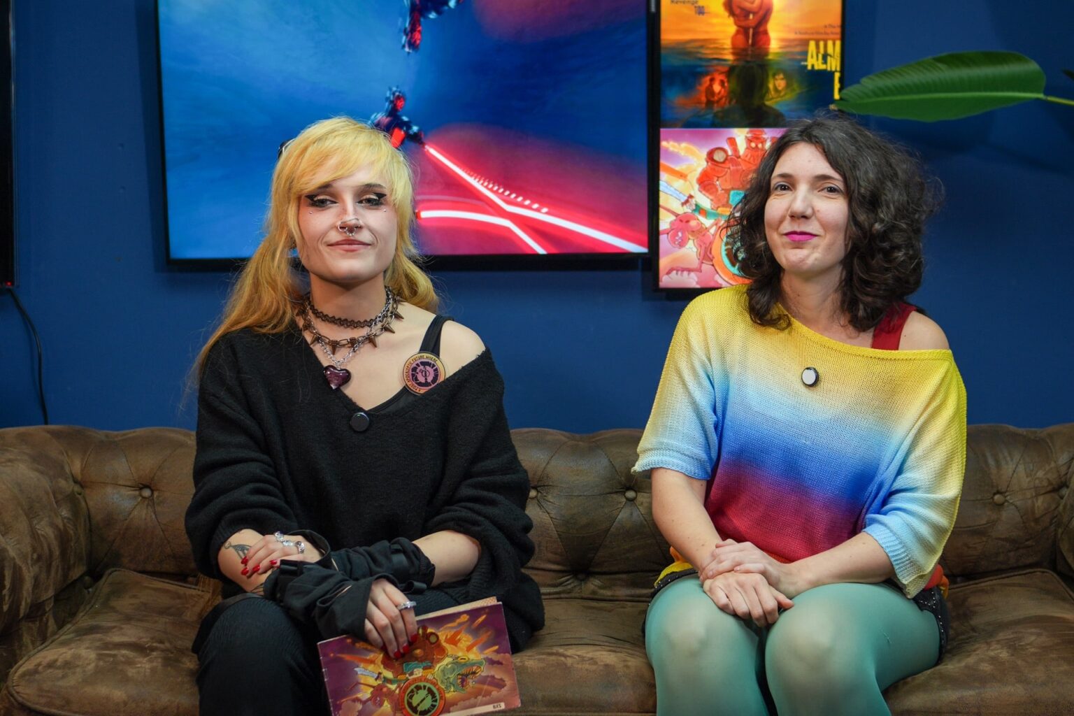 Ana morena con cabello ondulado y Yix rubia con pelo largo, sentadas en un sofá marrón en el espacio azul de la escuela, libros y pósters al fondo.