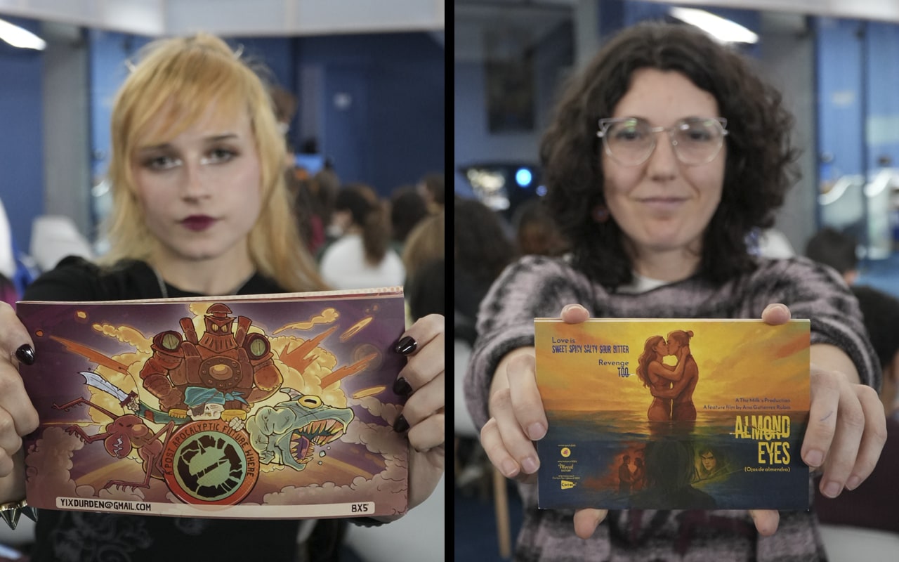 Ana y Yix muestran delante de ellas dos tarjetones con la portada de sus trabajos de animación en una sala con gente al fondo
