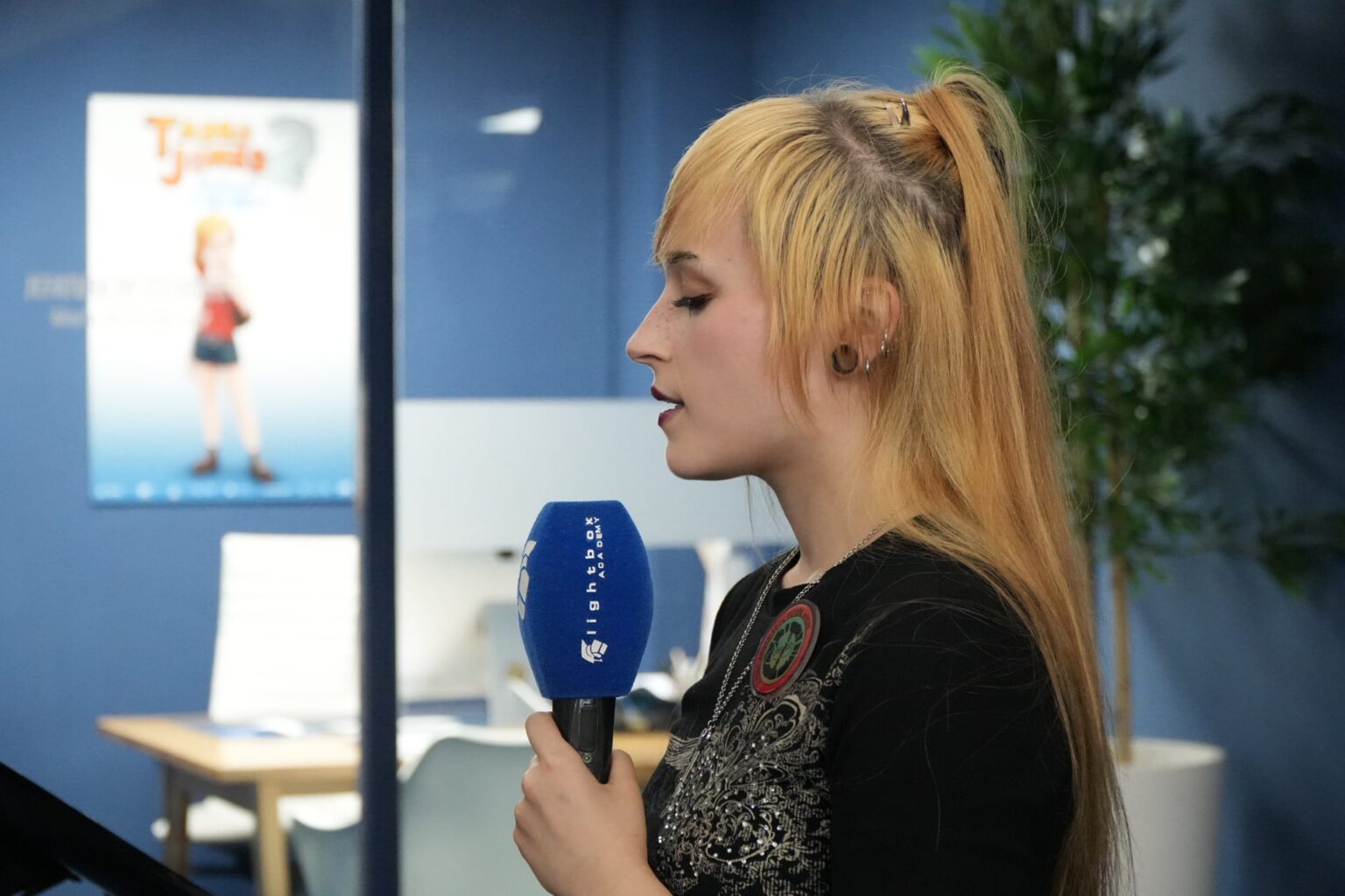 Yix, Mujer rubia con coleta alta sosteniendo un micrófono azul, perfil contra un póster de película animada en Lightbox Academy